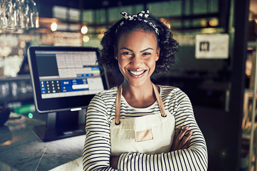 Smiling young African waitress wearing an apron and smiling while standing by a point of sale terminal in a trendy restaurant